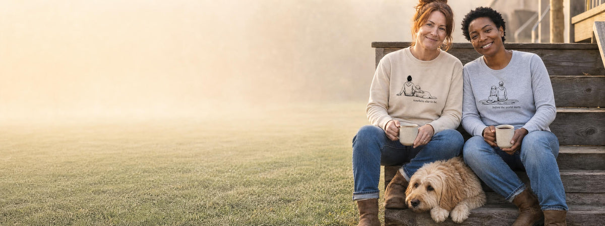 Two women on misty porch steps with coffee and a golden dog, wearing a Nowhere Else To Be crewneck and a Before The World Starts long sleeve