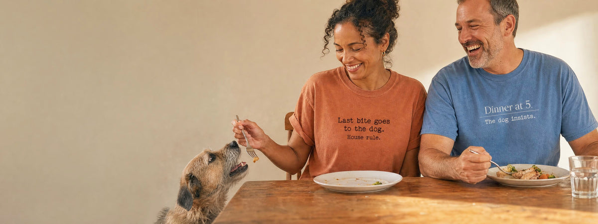 A woman in a Last Bite Goes to the Dog tee offers the last bite of dinner to her scruffy dog while a man in a Dinner at 5 tee laughs