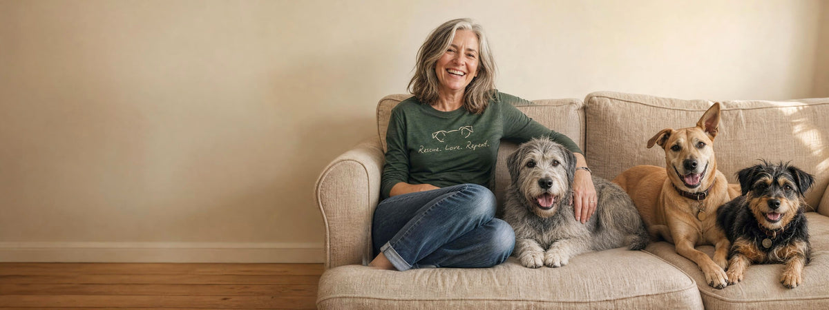 A woman in a Rescue Love Repeat long sleeve sits squeezed into the corner of a sofa with three rescue dogs