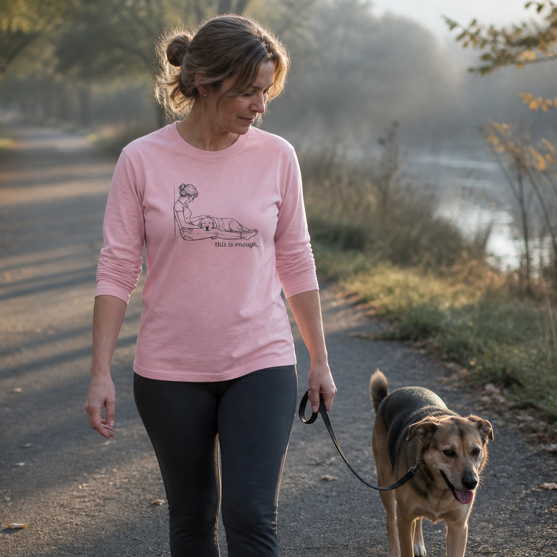 A woman walking a dog on a path outdoors, wearing a light pink long-sleeve graphic T-shirt and dark leggings. The shirt features a simple line drawing of a woman lying down with the text 'Mental Health Matters' below it.